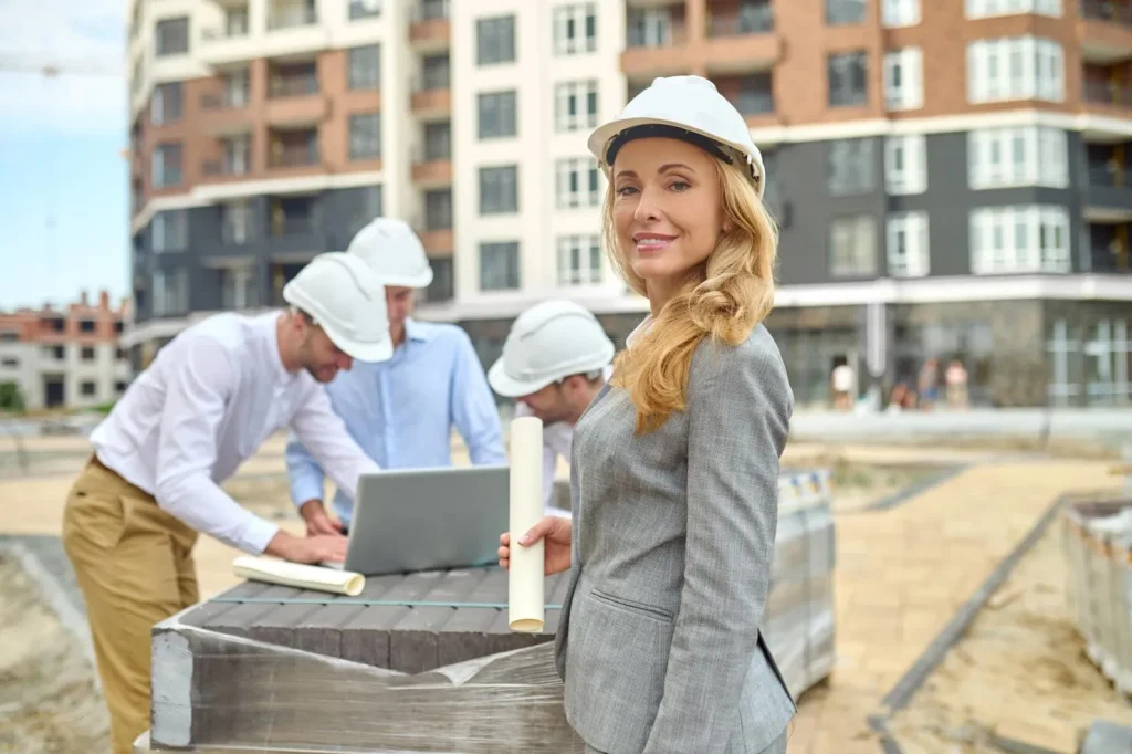 Female construction engineer wearing a white safety helmet holding building plans at a residential construction site with her team in the background.