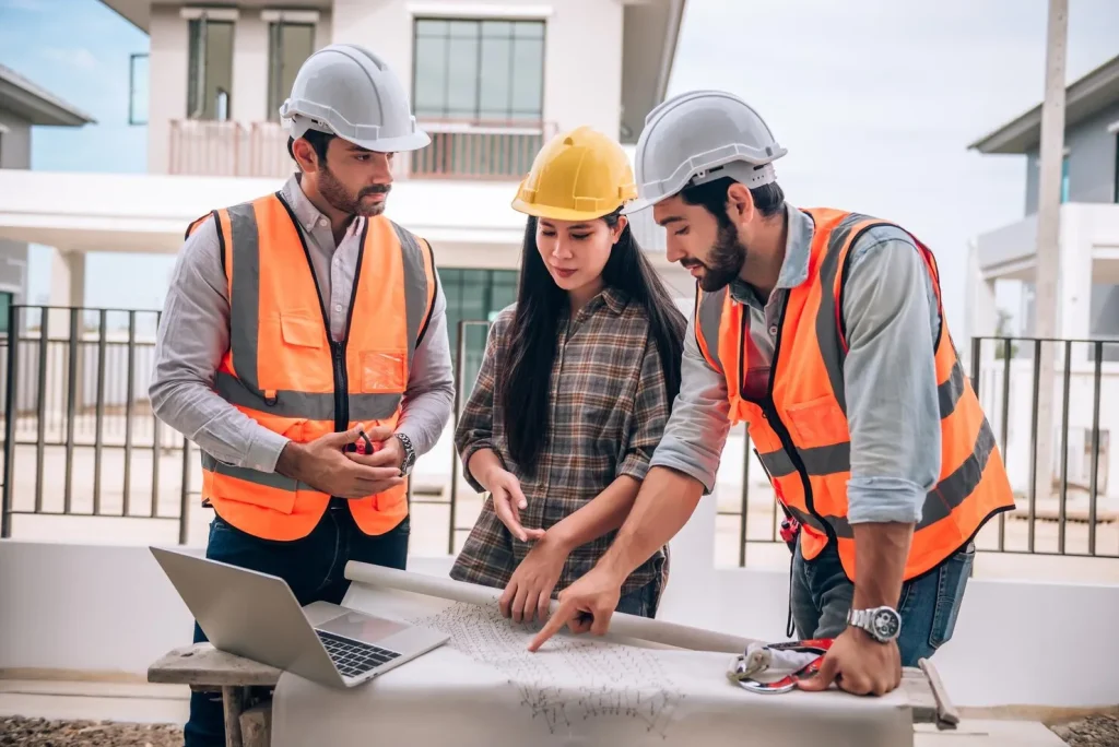 Three construction professionals wearing hard hats and high-visibility vests review architectural plans beside a laptop at a residential building site.